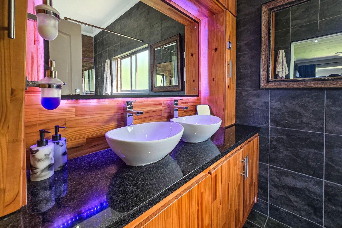 Modern bathroom featuring a double vanity with vessel sinks, granite countertop, wooden cabinetry, and stylish dark tiled walls with integrated LED lighting.