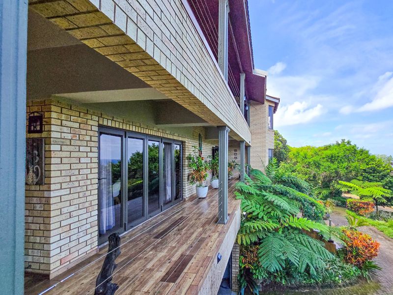 A spacious elevated balcony featuring timber-look decking, brick exterior walls, and sliding glass doors, overlooking a lush garden area.