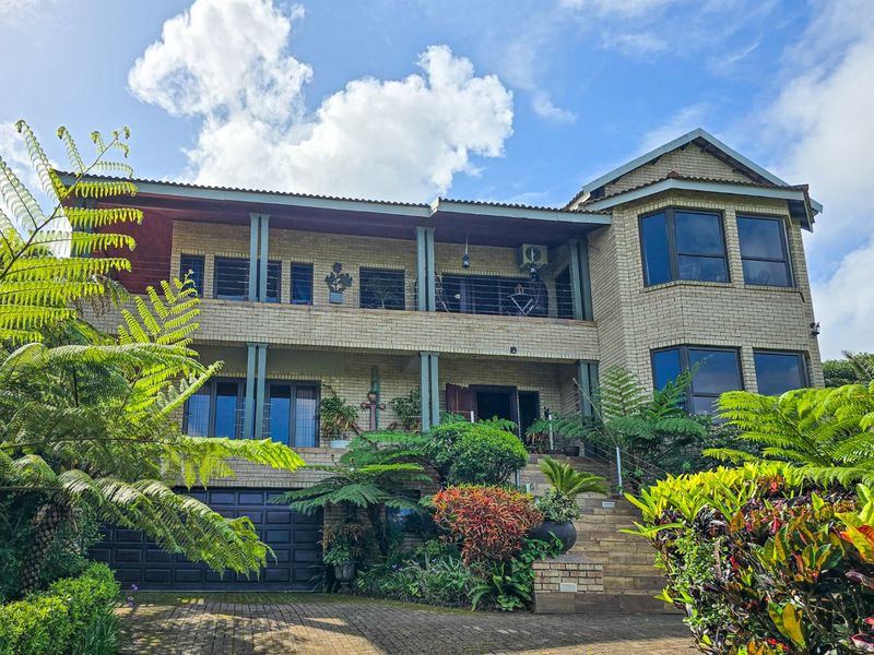 A double-story brick residence featuring a paved driveway, integrated garage, and a spacious upper-level balcony surrounded by established garden landscaping.