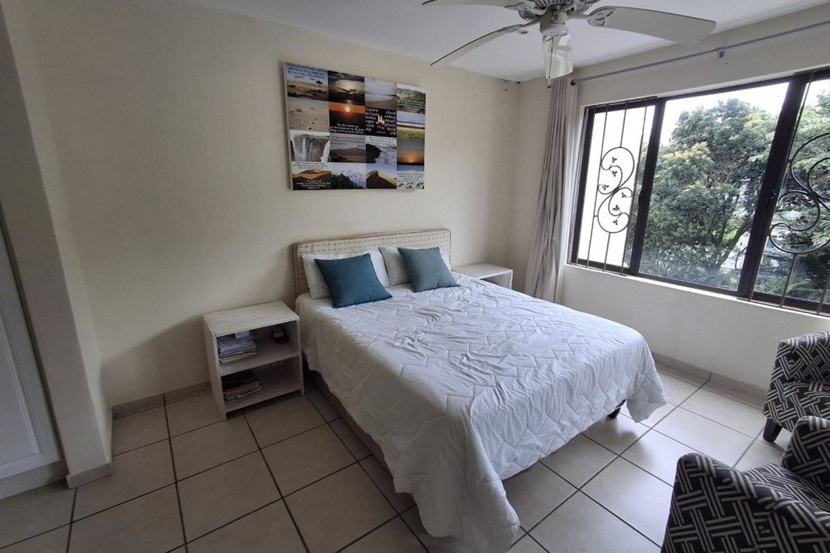 A bedroom featuring tiled flooring, a ceiling fan, and large windows with security bars, offering a functional and well-lit living space.