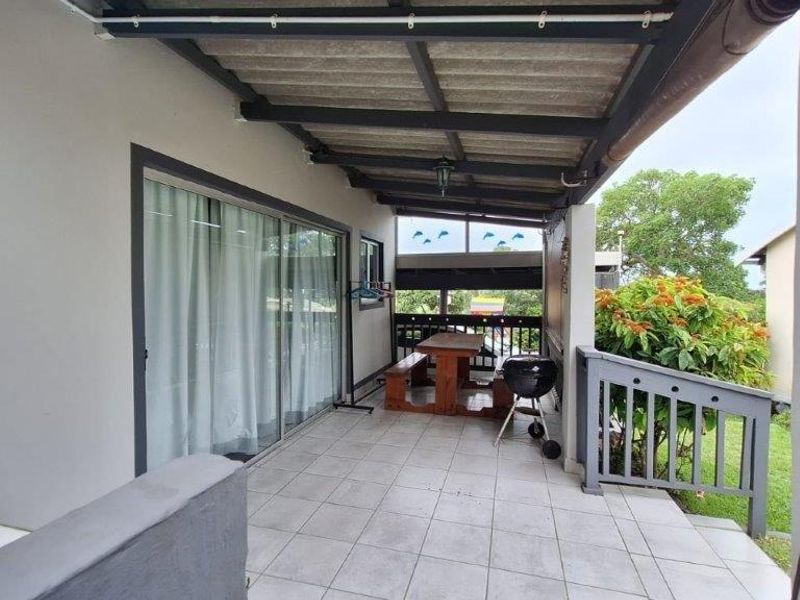 A covered patio area featuring tiled flooring, sliding glass door access, and a protective roof structure, overlooking the garden.