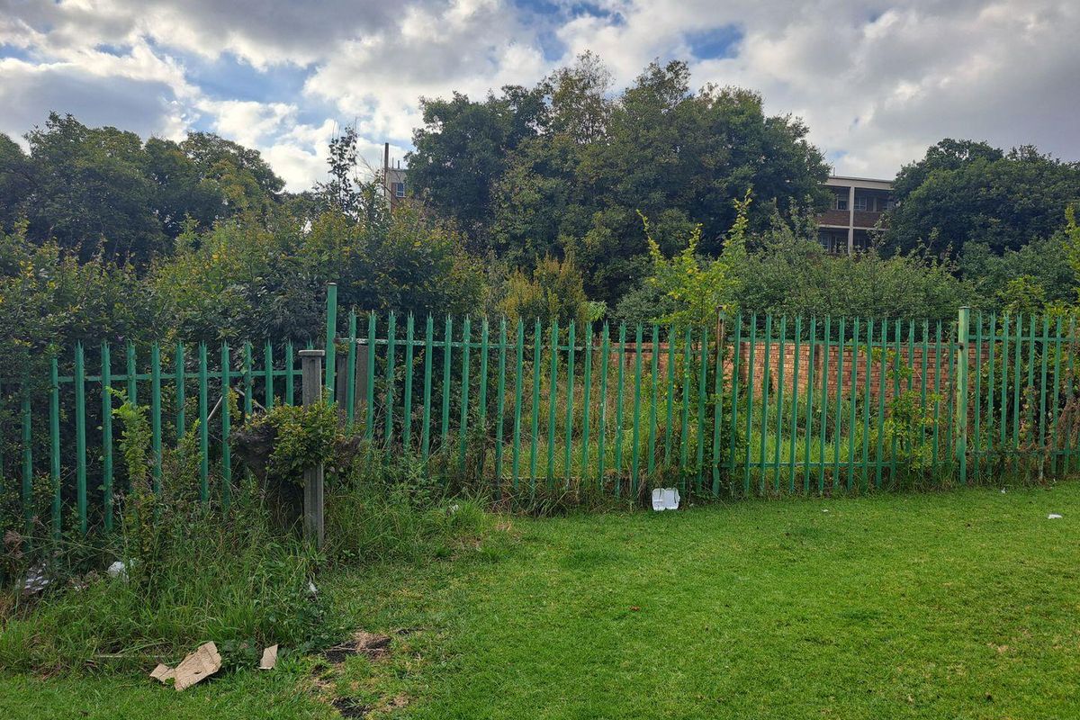 A view of the rear boundary featuring green palisade fencing, a brick wall, and mature trees on a residential plot.