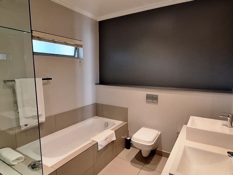 Modern bathroom featuring a double vanity, wall-mounted toilet, and a combined bathtub and glass-enclosed shower area with neutral tiling.
