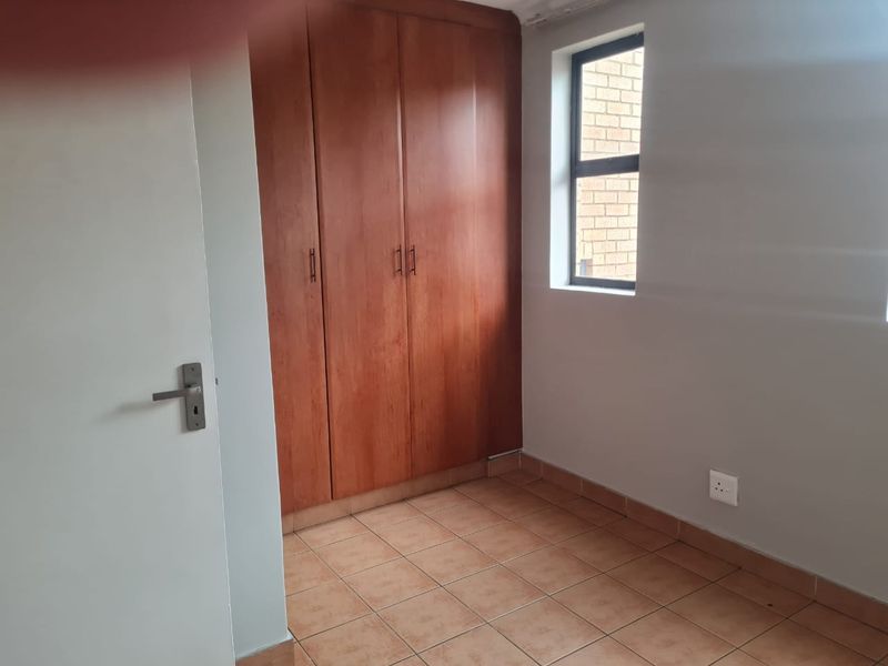 A compact, empty bedroom featuring tiled flooring, a built-in wooden wardrobe, and a window providing natural light.