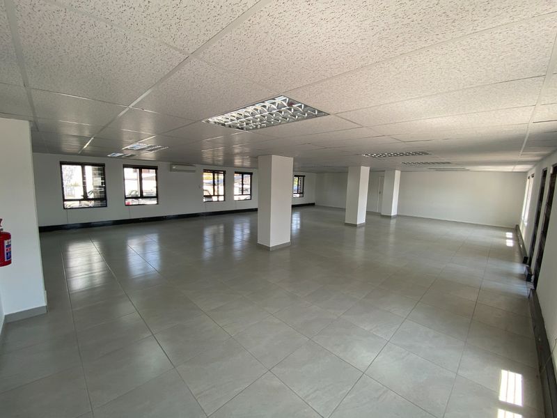 A spacious, open-plan commercial office area featuring neutral ceramic floor tiles, a suspended ceiling with recessed lighting, and structural support columns.
