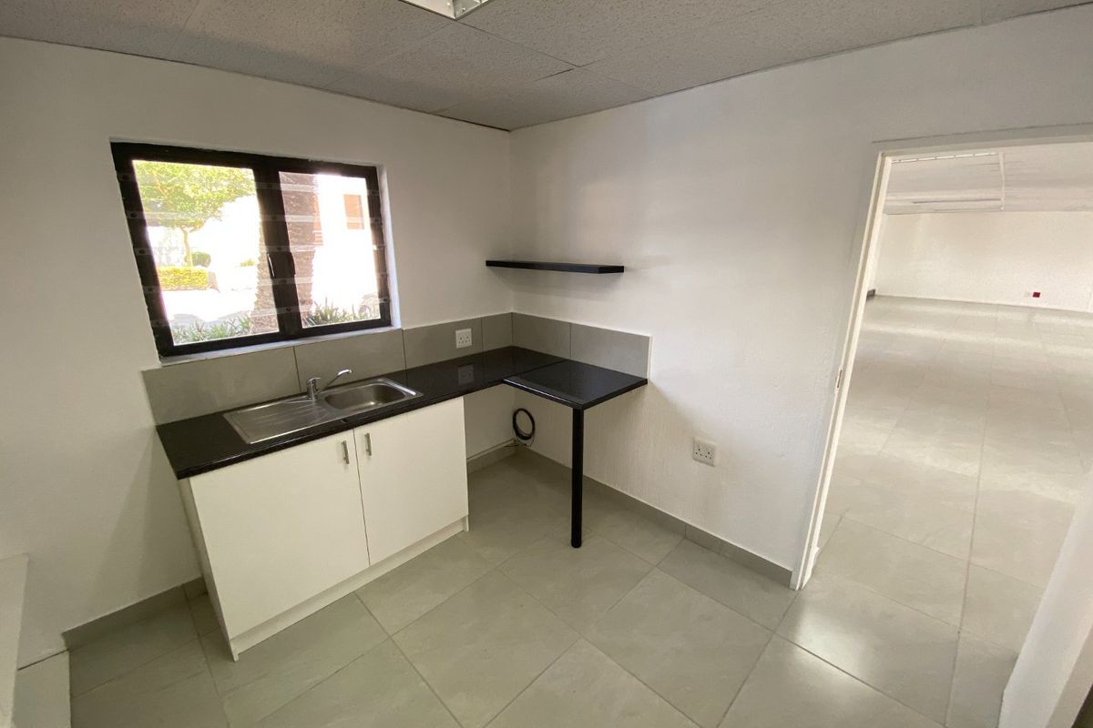 A compact kitchenette area featuring a stainless steel sink, dark laminate countertop, white cabinetry, and tiled flooring within a commercial office space.