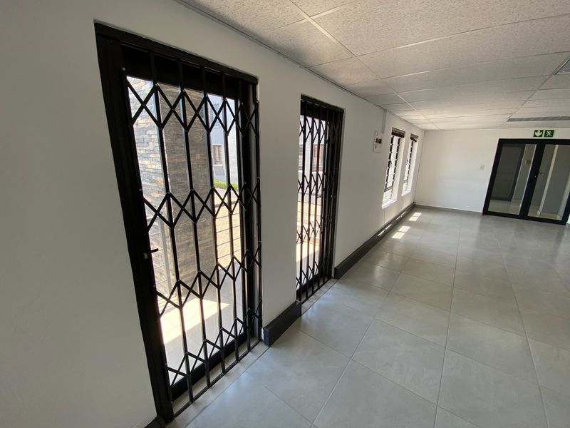 A bright, empty commercial hallway featuring tiled flooring, a suspended ceiling, and secure metal security gates leading to an outdoor area.