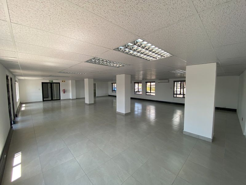 A spacious, open-plan commercial office area featuring neutral ceramic tiling, a suspended ceiling with recessed lighting, and large windows providing ample natural light.