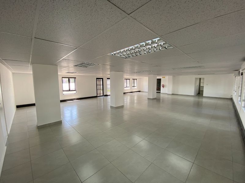 A spacious, open-plan commercial office area featuring neutral ceramic floor tiles, a suspended ceiling with recessed lighting, and multiple structural support columns.