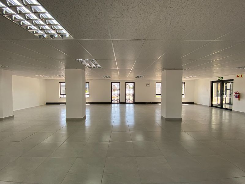A spacious, open-plan commercial office area featuring neutral ceramic floor tiles, a suspended ceiling with recessed lighting, and large windows with security gates.