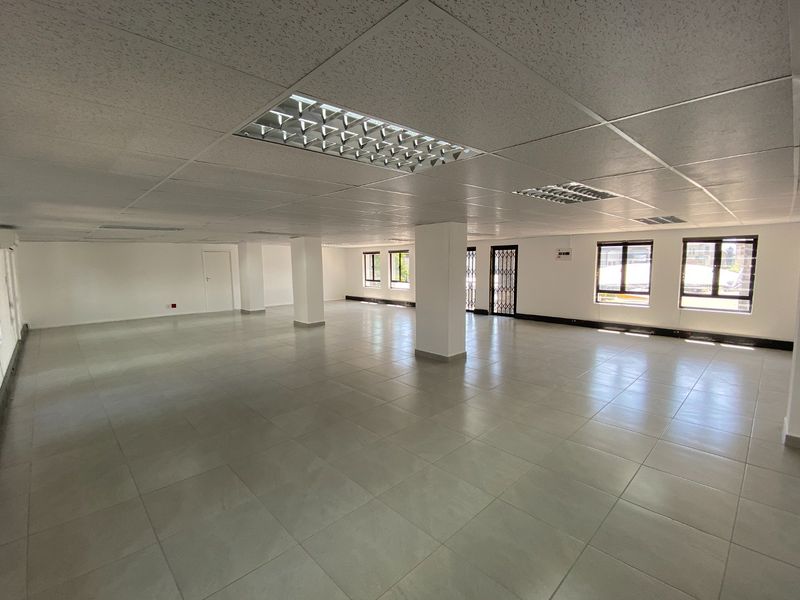A spacious, open-plan commercial office area featuring neutral ceramic floor tiles, a suspended ceiling with recessed lighting, and large windows providing ample natural light.