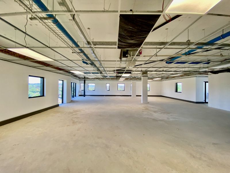 A large, open-plan commercial space in shell condition, featuring exposed ceiling infrastructure, concrete floors, and multiple windows providing natural light.