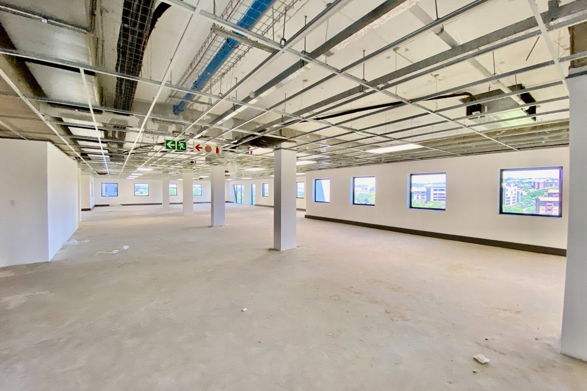 A large, open-plan commercial office space in shell condition, featuring exposed ceiling infrastructure, structural columns, and large windows providing natural light.