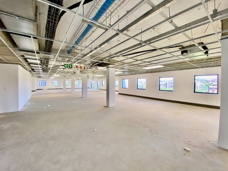 A large, open-plan commercial office space in shell condition, featuring exposed ceiling infrastructure, structural columns, and large windows providing natural light.