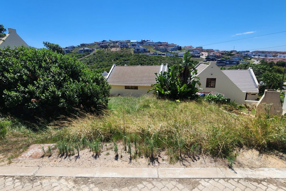 A vacant residential plot featuring a sloped landscape with established surrounding vegetation and views of the neighboring residential area.