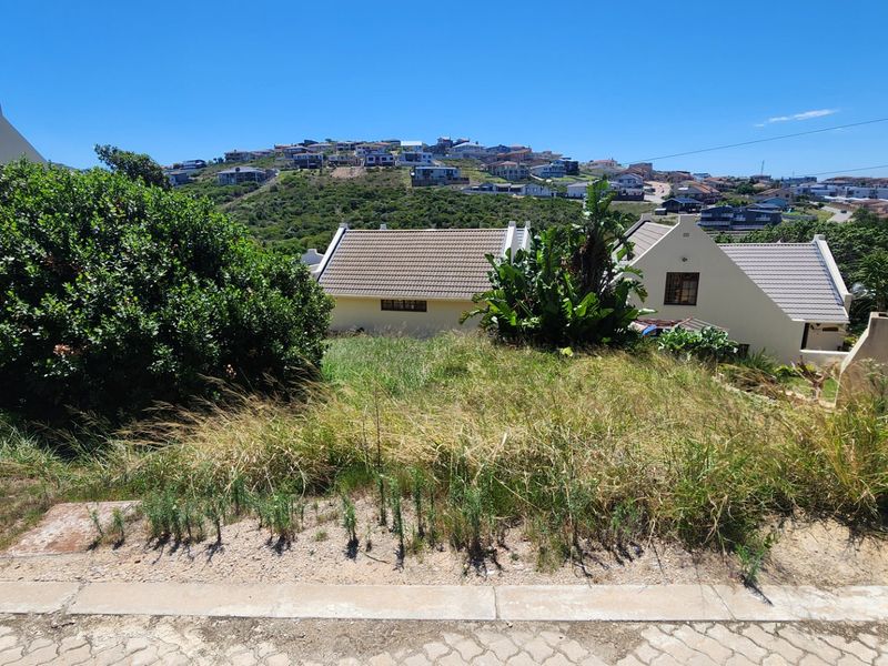 A vacant residential plot featuring a sloped landscape with established surrounding vegetation and views of the neighboring residential area.