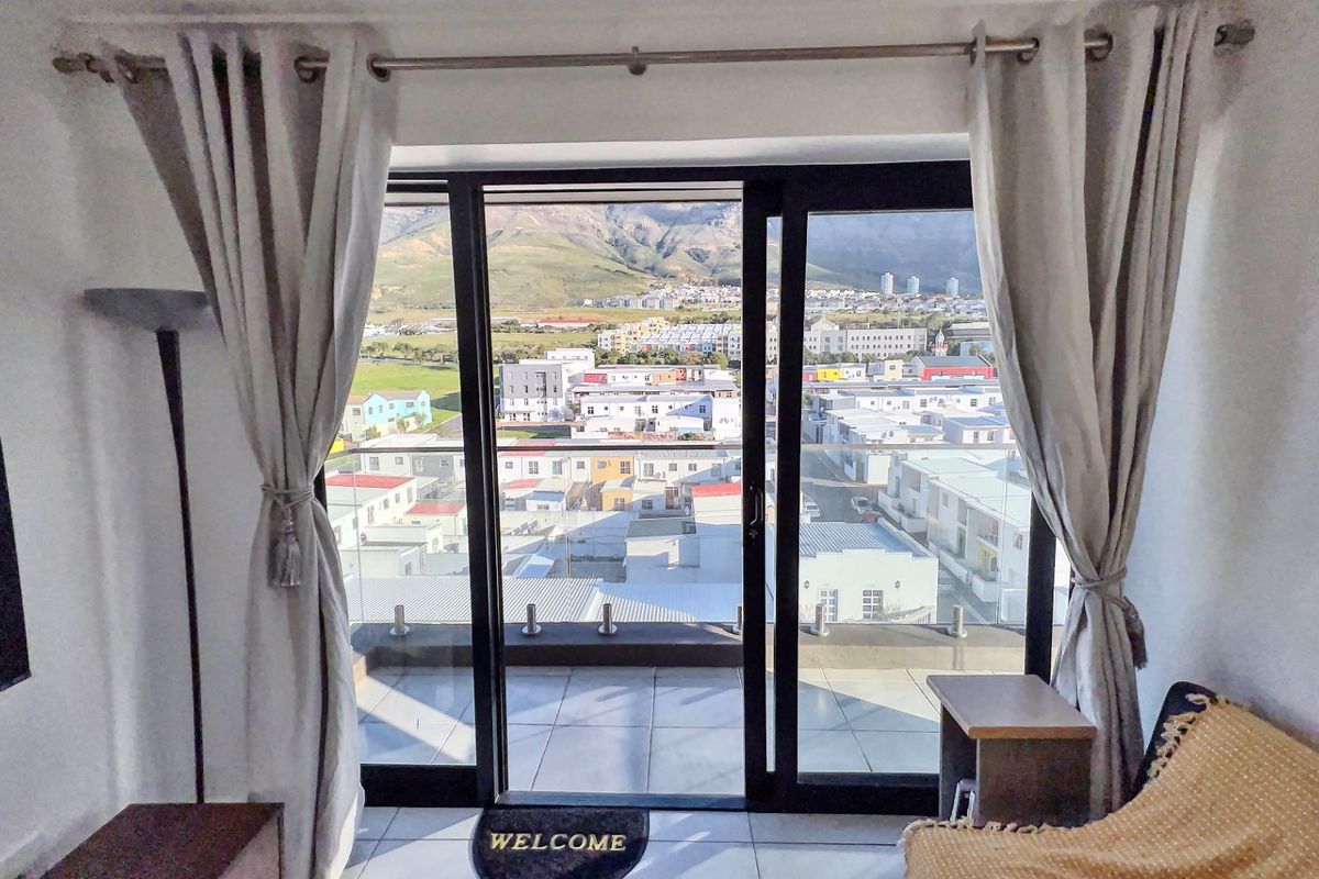 A bright living area featuring large sliding glass doors that open onto a balcony with a glass balustrade and expansive mountain and urban views.