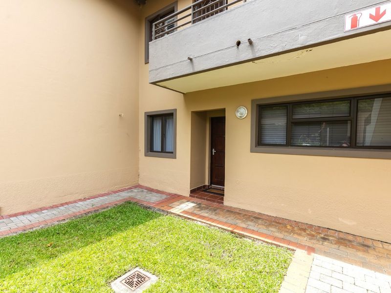 A ground floor apartment exterior featuring a small private lawn, paved walkway, and an overhead balcony with metal railings.