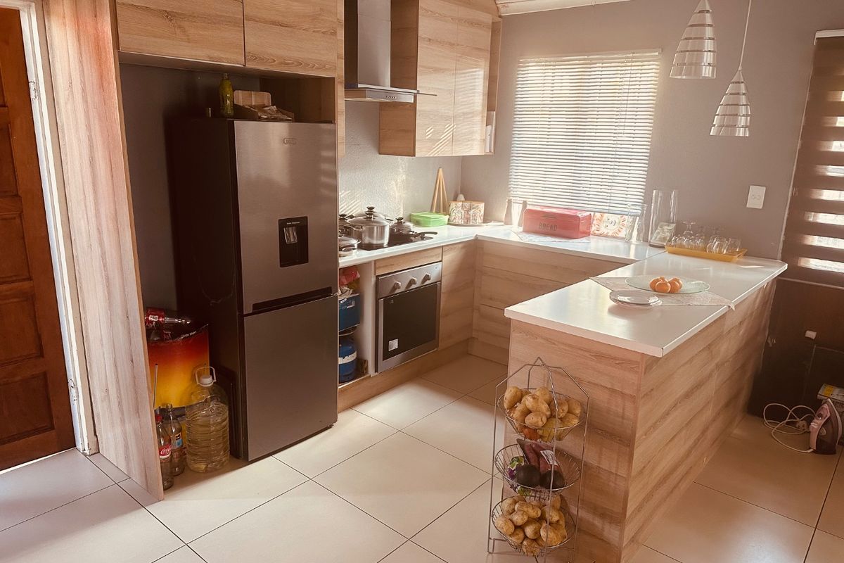 A modern kitchen featuring light wood-grain cabinetry, stone countertops, a breakfast bar, and integrated appliances, complemented by neutral tiled flooring and natural light.