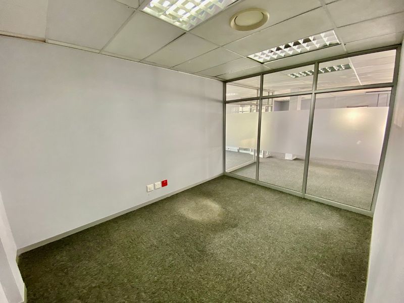 An empty commercial office space featuring a glass partition wall, suspended ceiling with recessed lighting, and carpeted flooring.