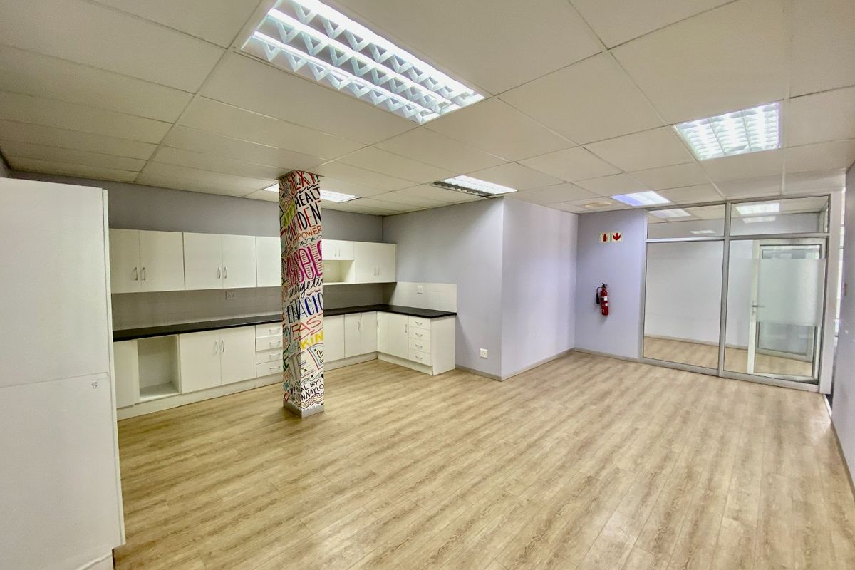 An open-plan commercial kitchen area featuring laminate flooring, built-in cabinetry with dark countertops, drop ceilings, and a glass partition wall.