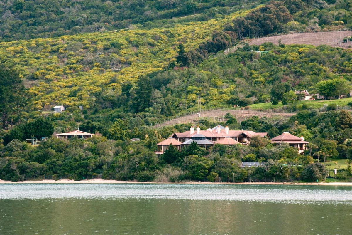 A scenic view of a large estate property nestled against a lush, forested hillside overlooking the water.