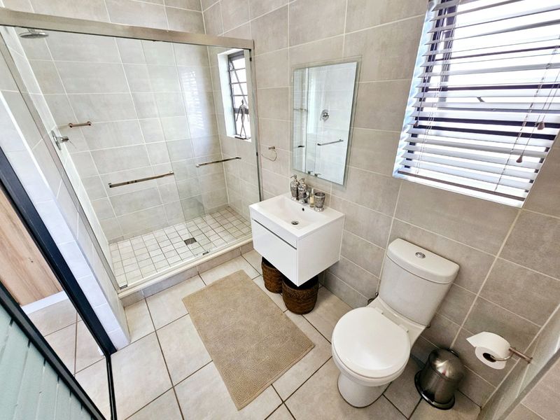 Modern bathroom featuring a glass-enclosed shower, wall-mounted vanity with basin, toilet, and neutral floor-to-ceiling tiling with venetian blinds.