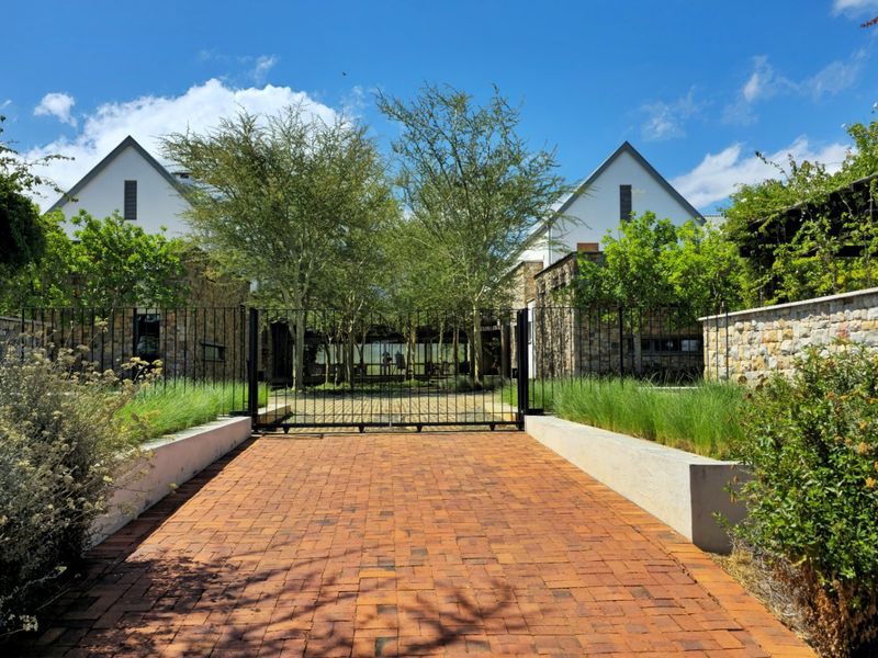 A well-maintained farm property entrance featuring a brick driveway, stone-clad building facades, and a secure metal gate set within landscaped grounds.