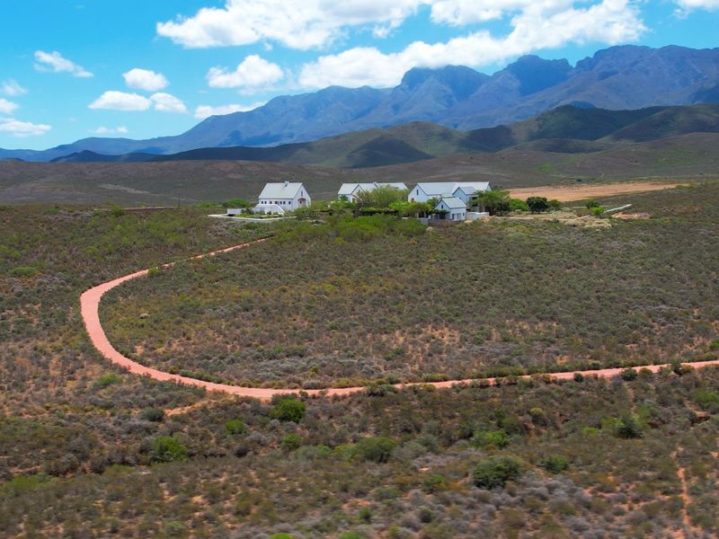 Aerial view of a farm property featuring multiple buildings with pitched roofs, set within a vast landscape with a long gravel driveway.