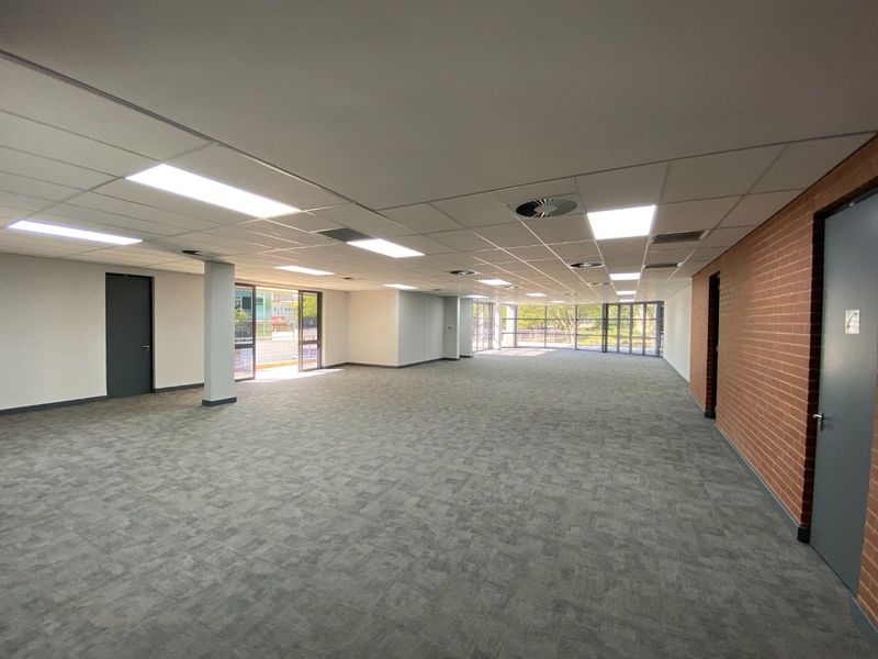 Spacious, open-plan commercial office area featuring carpeted flooring, suspended ceilings with recessed lighting, and large glass doors providing natural light.
