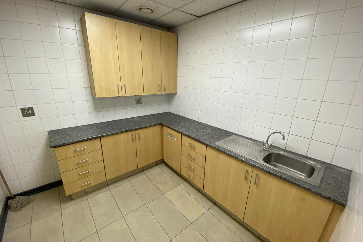 A functional commercial kitchenette featuring light wood-finish cabinetry, granite-look countertops, a stainless steel sink, and full-height wall tiling.