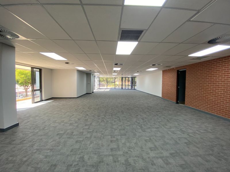 A spacious, open-plan commercial office area featuring carpeted flooring, a suspended ceiling with recessed lighting, and an accent brick wall with large windows.