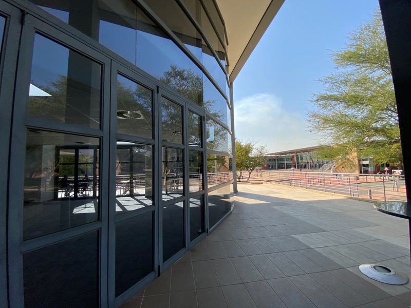 A spacious covered outdoor patio area featuring large glass facade panels, tiled flooring, and metal safety railings overlooking the commercial property grounds.
