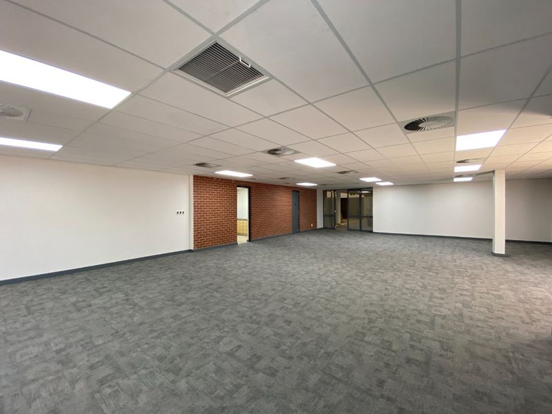 Spacious open-plan commercial office area featuring a suspended ceiling, recessed lighting, carpet tiles, and an exposed brick feature wall.