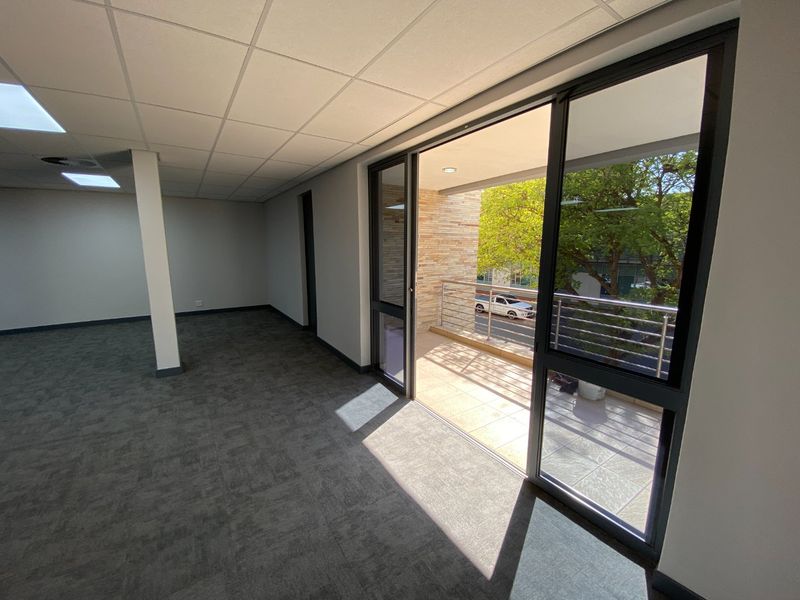 A bright, empty office space featuring carpet tiles, a suspended ceiling, and direct access to a private balcony with stainless steel railings.