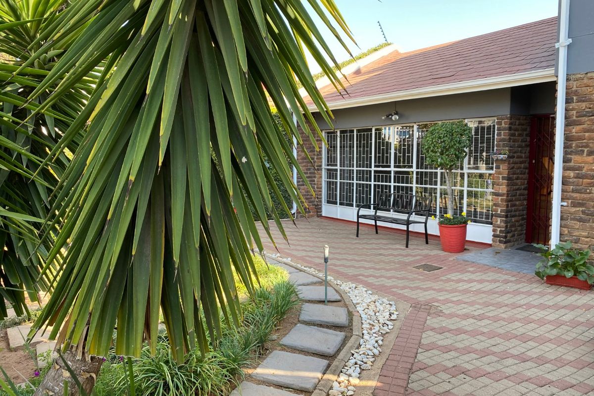 A paved outdoor patio area featuring a brick facade, security-barred windows, and a landscaped garden path leading to the entrance.