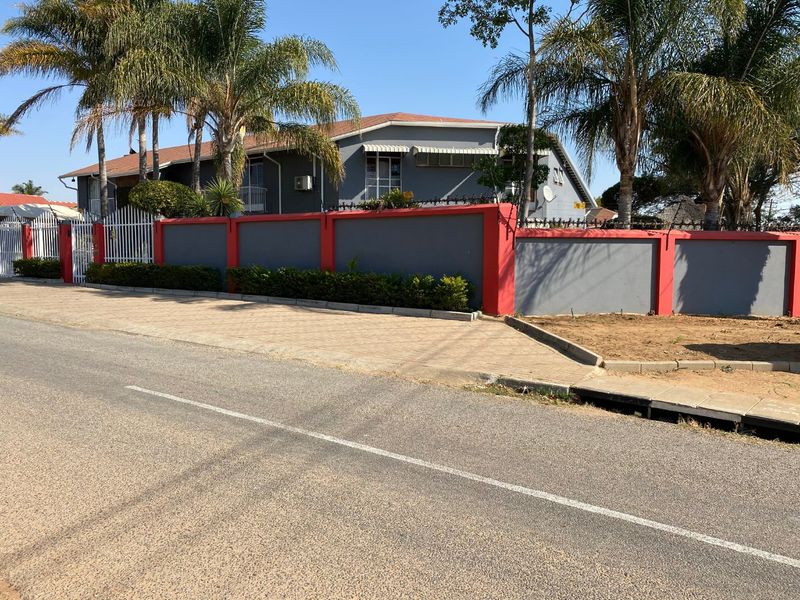 Front view of a residential property featuring a secure perimeter wall with metal gates, paved driveway, and established palm trees.