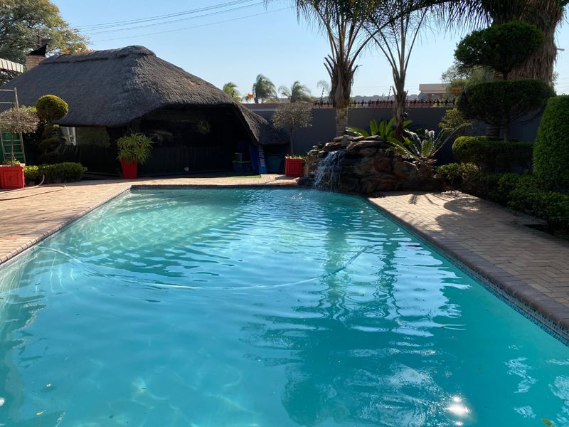 Outdoor pool area featuring a swimming pool with a rock water feature and an adjacent thatched lapa, set within a landscaped garden.