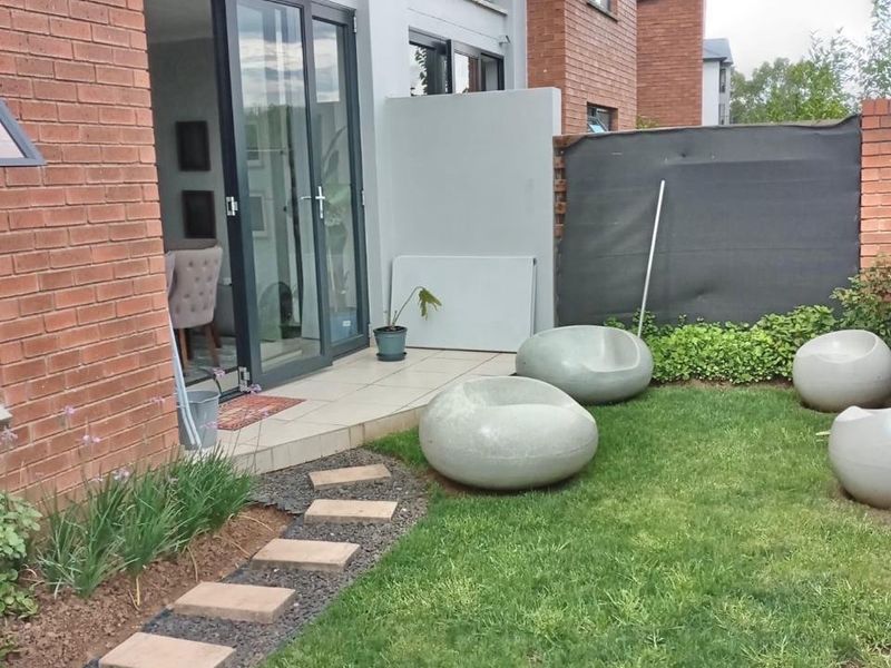 A private rear garden featuring a paved patio area, stone walkway, and lawn, accessible via glass sliding doors from the apartment.