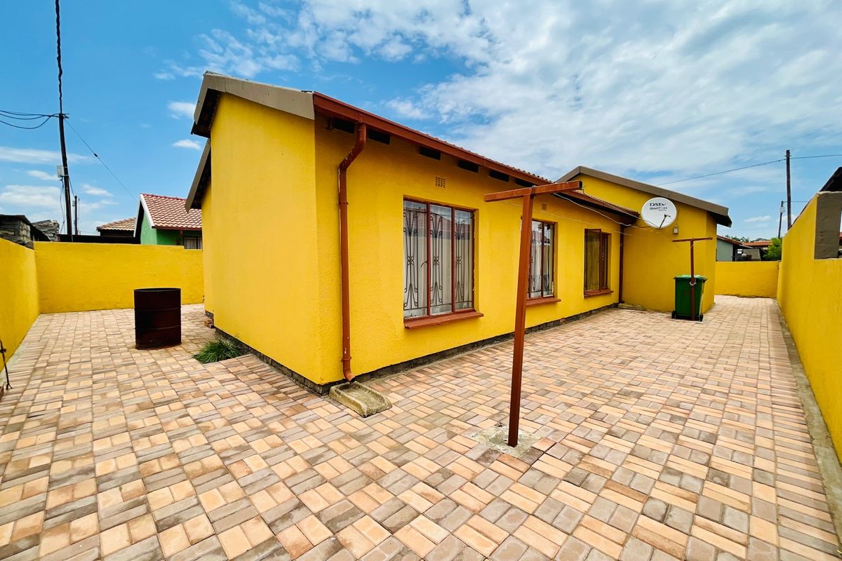 A view of the rear exterior of the house featuring paved courtyard area, yellow painted walls, and security bars on windows.