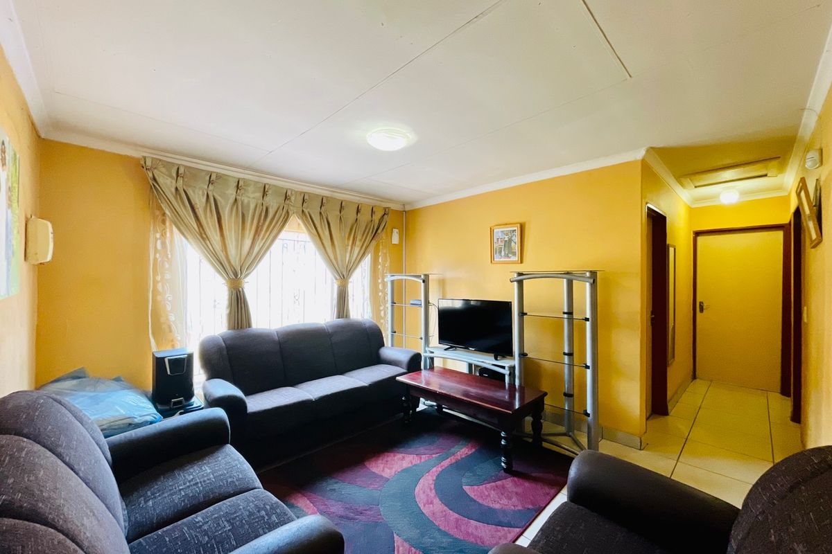 A living room featuring yellow walls, tiled flooring, and a window with curtain rails, providing access to an adjacent hallway.