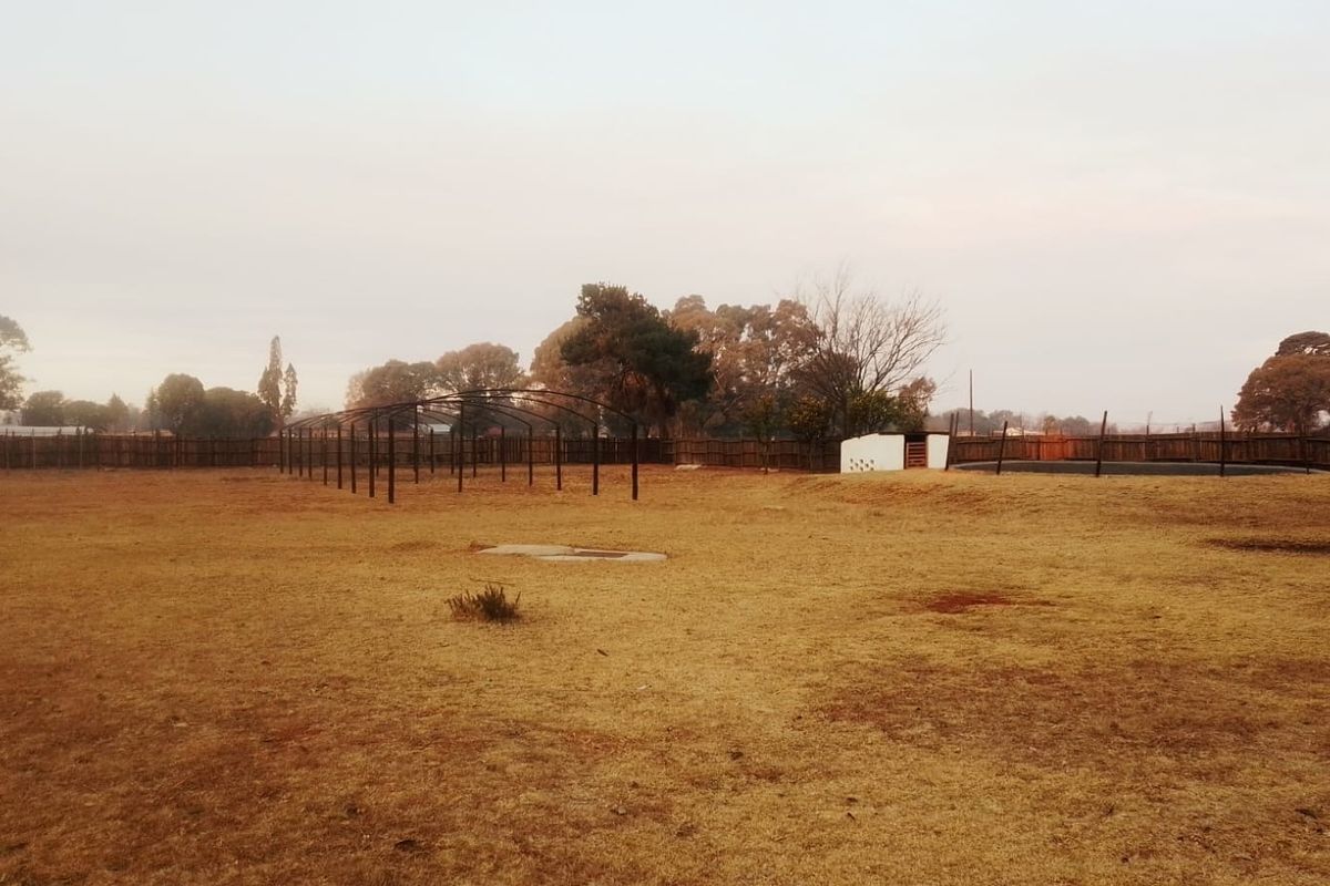 A large, open backyard area featuring a metal frame structure, perimeter wooden fencing, and a small masonry outbuilding on a dry, unlandscaped plot.