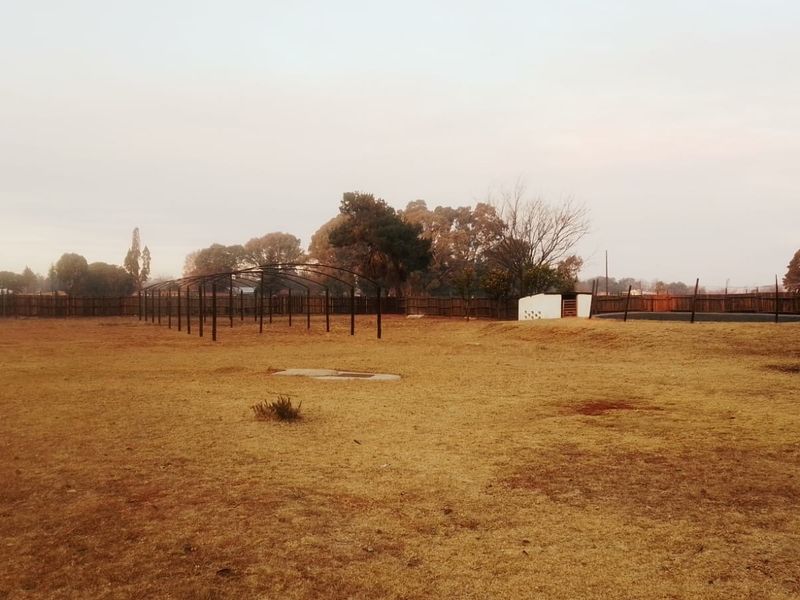 A large, open backyard area featuring a metal frame structure, perimeter wooden fencing, and a small masonry outbuilding on a dry, unlandscaped plot.