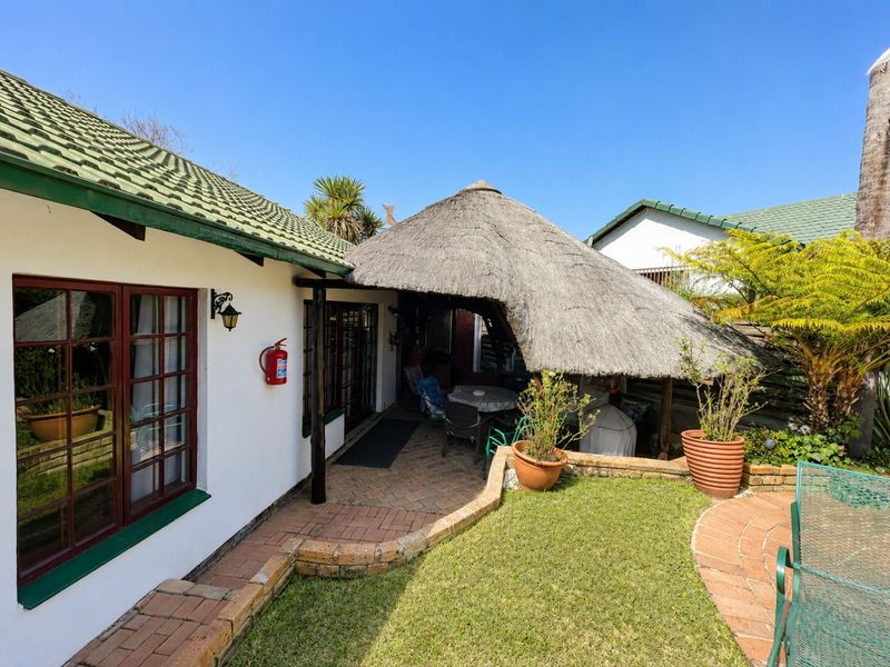 A covered outdoor patio area featuring a traditional thatched roof, brick paving, and a low brick retaining wall overlooking a small garden space.