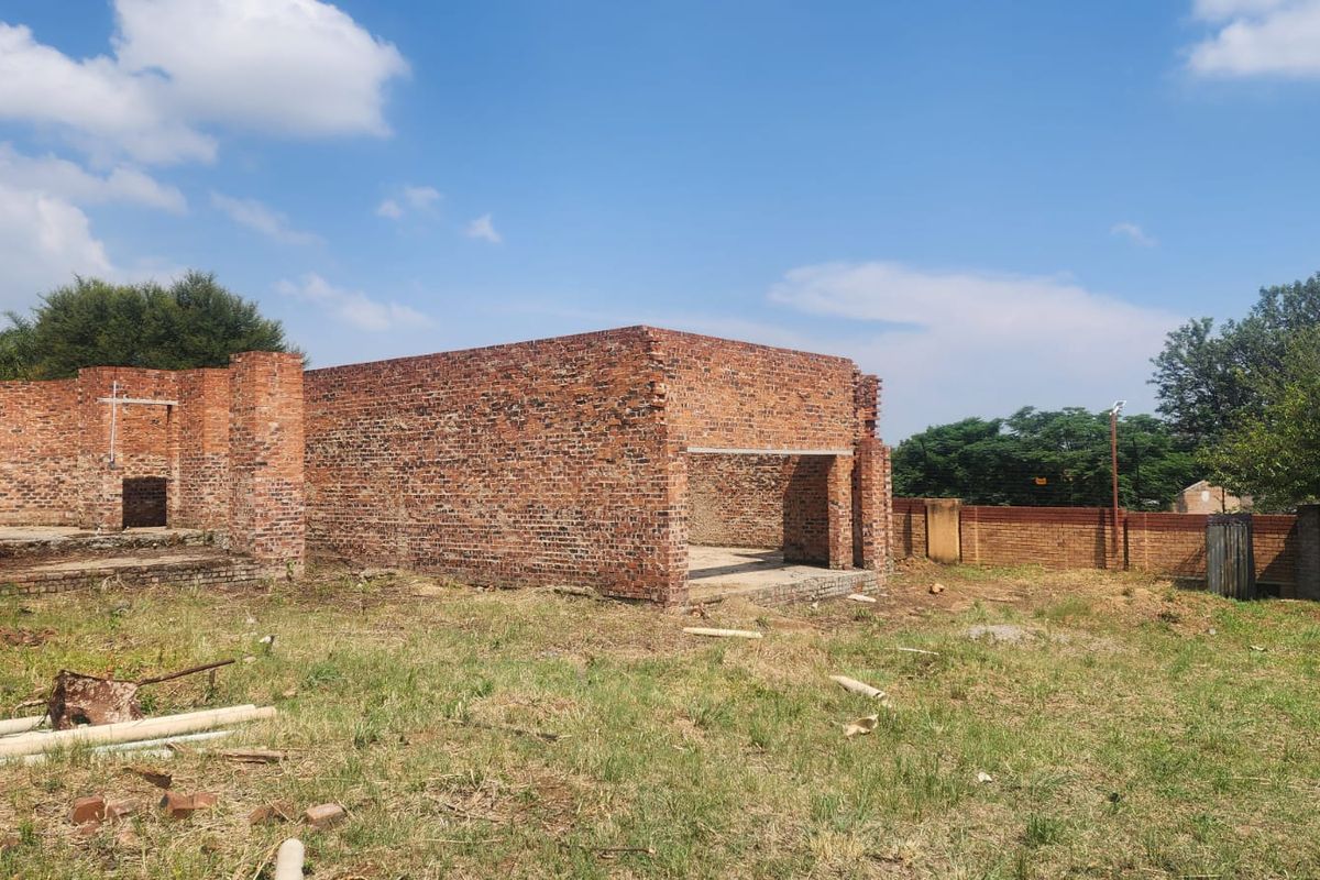An incomplete brick structure on a vacant residential plot, showing exposed masonry walls and concrete foundation work.