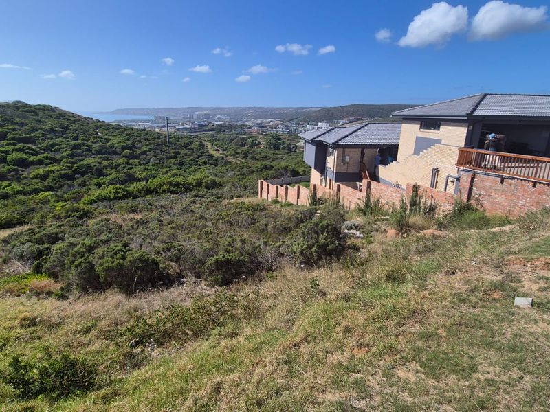 A view of a vacant residential plot featuring natural vegetation, with a neighboring property and distant coastal landscape in the background.