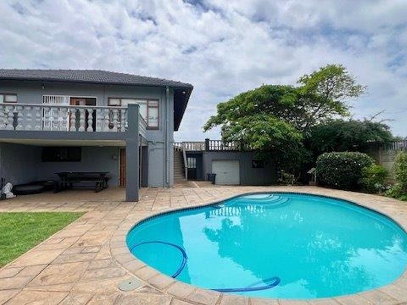 Rear exterior view featuring a swimming pool, paved patio area, elevated balcony with balustrades, and access to the garage and garden.