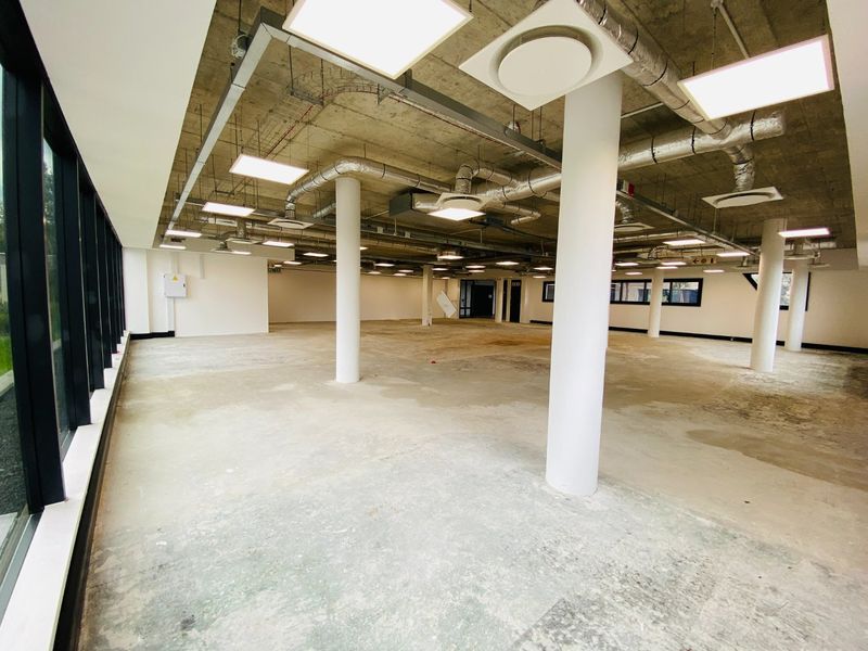 Spacious open-plan commercial office area featuring an industrial aesthetic with exposed concrete ceilings, HVAC ducting, and large windows providing ample natural light.