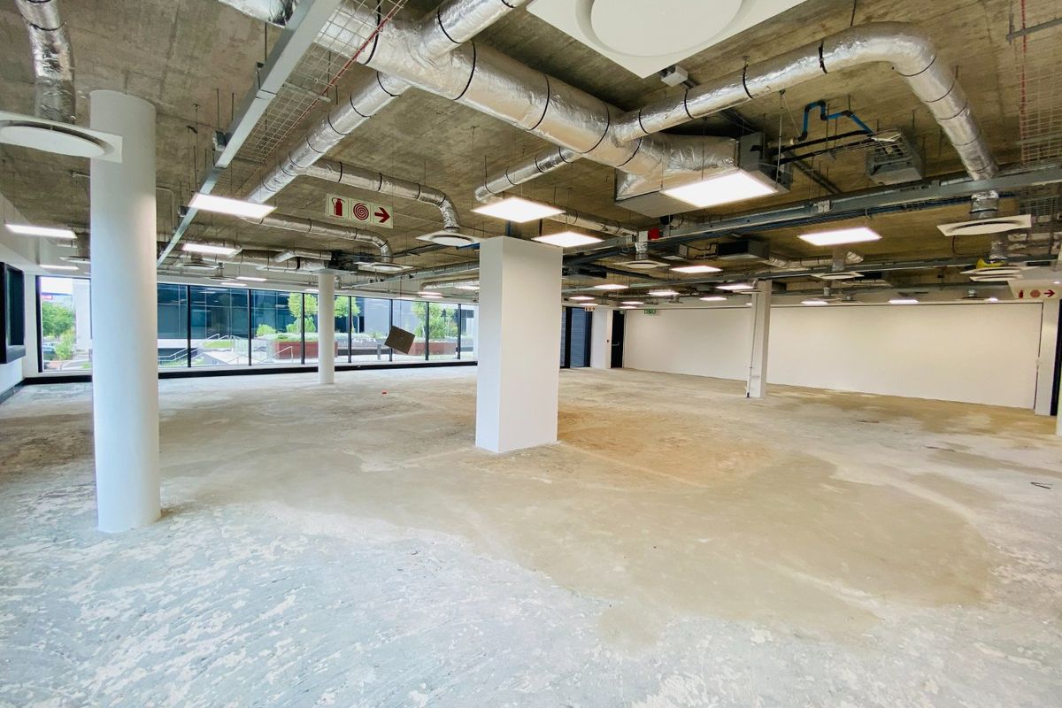 Spacious open-plan commercial office area featuring an industrial aesthetic with exposed concrete ceilings, HVAC ducting, and large windows providing ample natural light.