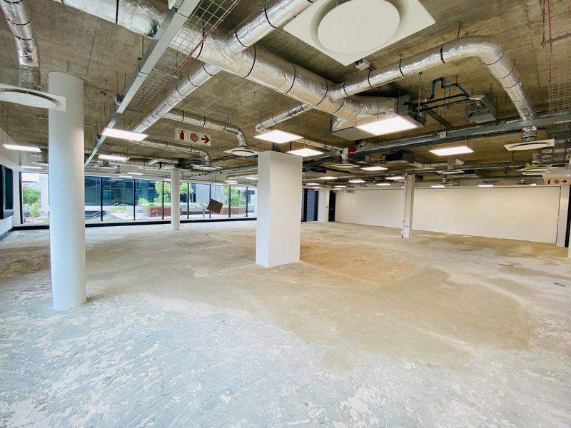 Spacious open-plan commercial office area featuring an industrial aesthetic with exposed concrete ceilings, HVAC ducting, and large windows providing ample natural light.